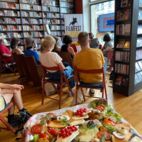 Im Vordergrund ein Tablet mit Schnittchen und Snacks, im Hintergrund eine sitzende Gruppe von Menschen